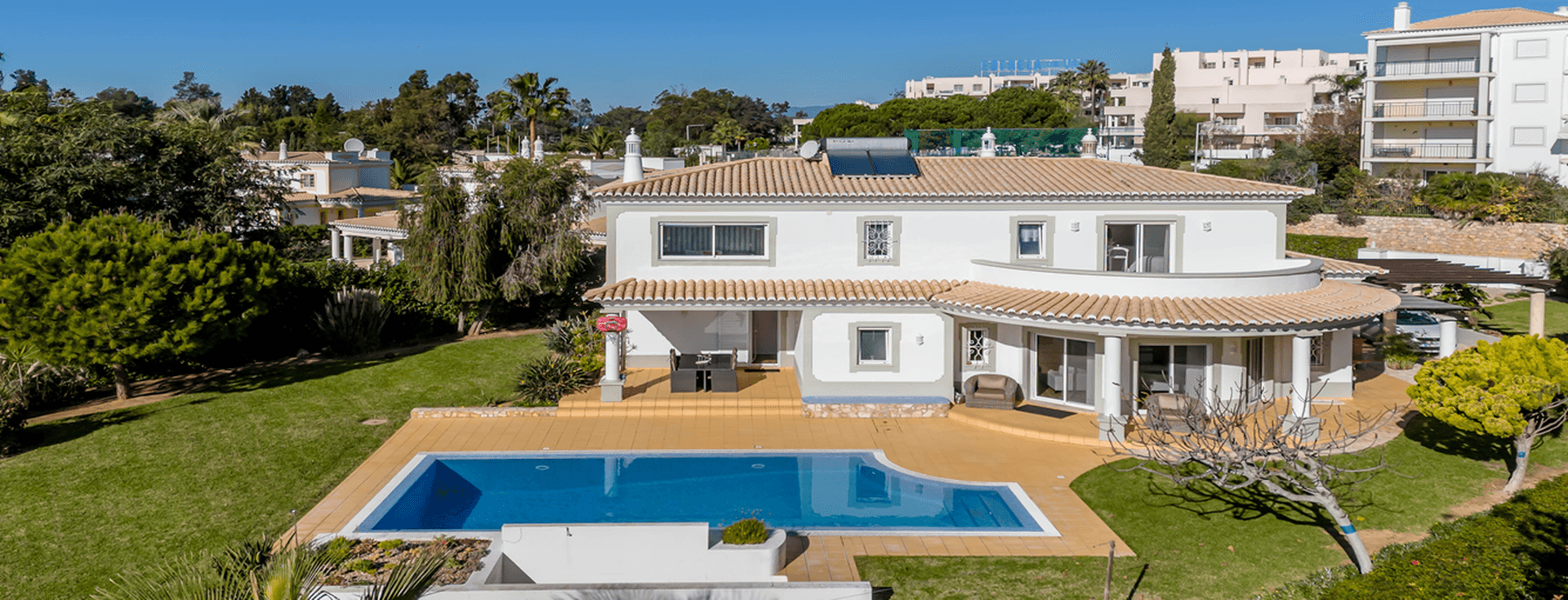 Luftaufnahme einer modernen weißen Villa mit Ziegeldach, großem Pool und üppigem Garten, gelegen in einem Wohngebiet mit klarem blauen Himmel.