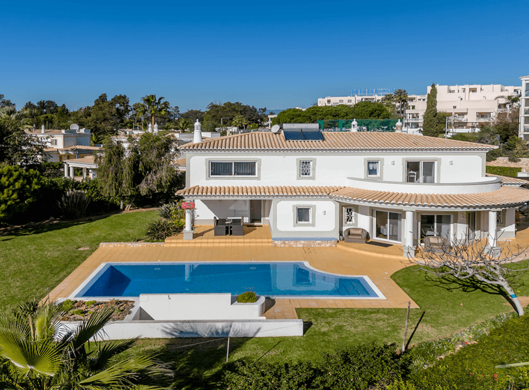 Luftaufnahme einer modernen weißen Villa mit Ziegeldach, großem Pool und üppigem Garten, gelegen in einem Wohngebiet mit klarem blauen Himmel.