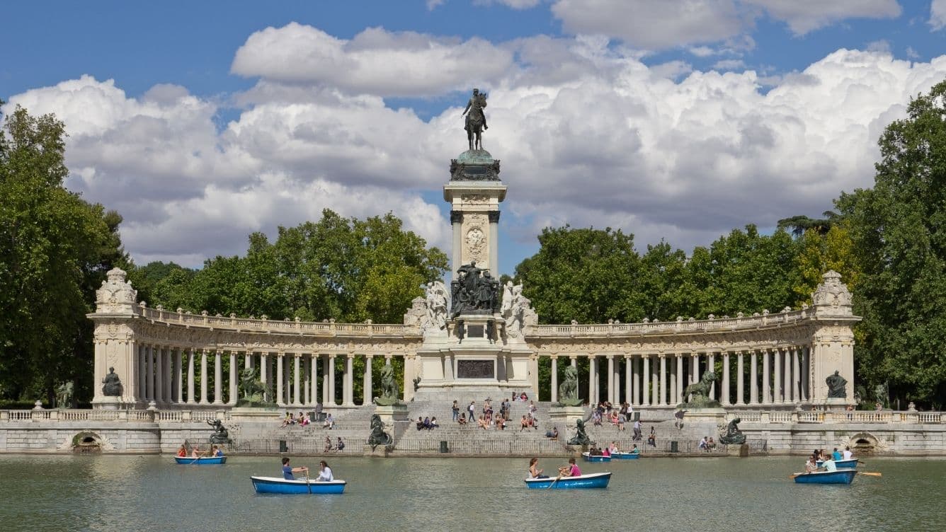 People rowing boats on a lake in front of the Monument to Alfonso XII in Madrid's Retiro Park with a backdrop of trees and clouds.