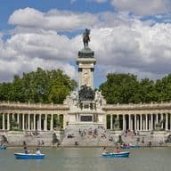 Gente remando en barcas en un lago frente al Monumento a Alfonso XII en el Parque del Retiro de Madrid, con un telón de fondo de árboles y nubes.