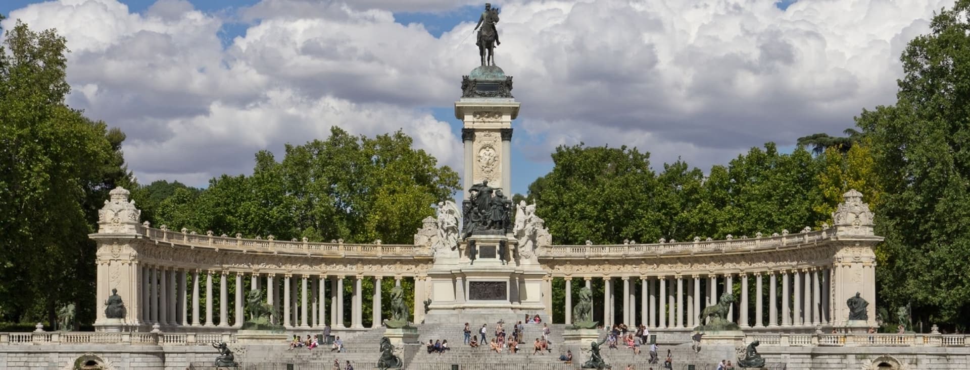 People rowing boats on a lake in front of the Monument to Alfonso XII in Madrid's Retiro Park with a backdrop of trees and clouds.