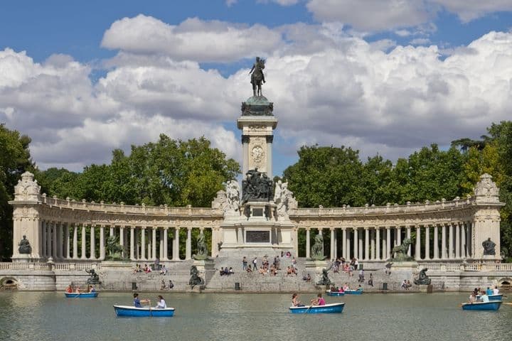 Gente remando en barcas en un lago frente al Monumento a Alfonso XII en el Parque del Retiro de Madrid, con un telón de fondo de árboles y nubes.