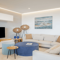 Modernes Wohnzimmer mit einer beigen Eckcouch, einem blauen Ottoman, Flachbildfernseher, abstrakter Wandkunst und Korbstehlampen auf einem hellen Teppich.