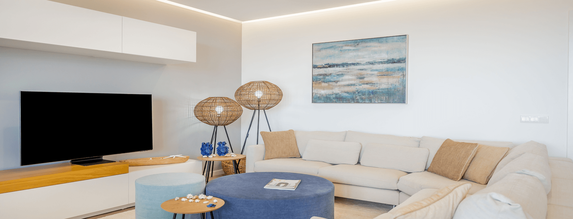 Sala de estar moderna com sofá seccional bege, pufe azul, televisão de ecrã plano, arte abstrata na parede e candeeiros de chão em verga sobre um tapete claro.