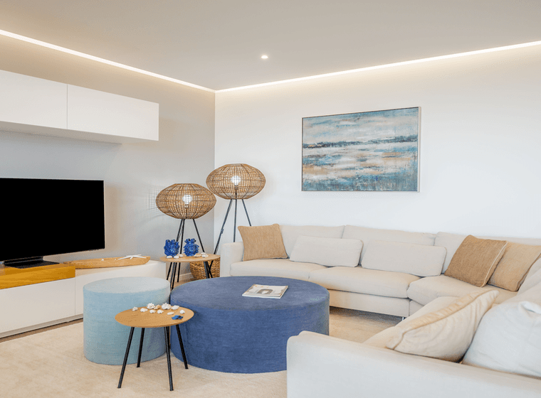 Modernes Wohnzimmer mit einer beigen Eckcouch, einem blauen Ottoman, Flachbildfernseher, abstrakter Wandkunst und Korbstehlampen auf einem hellen Teppich.