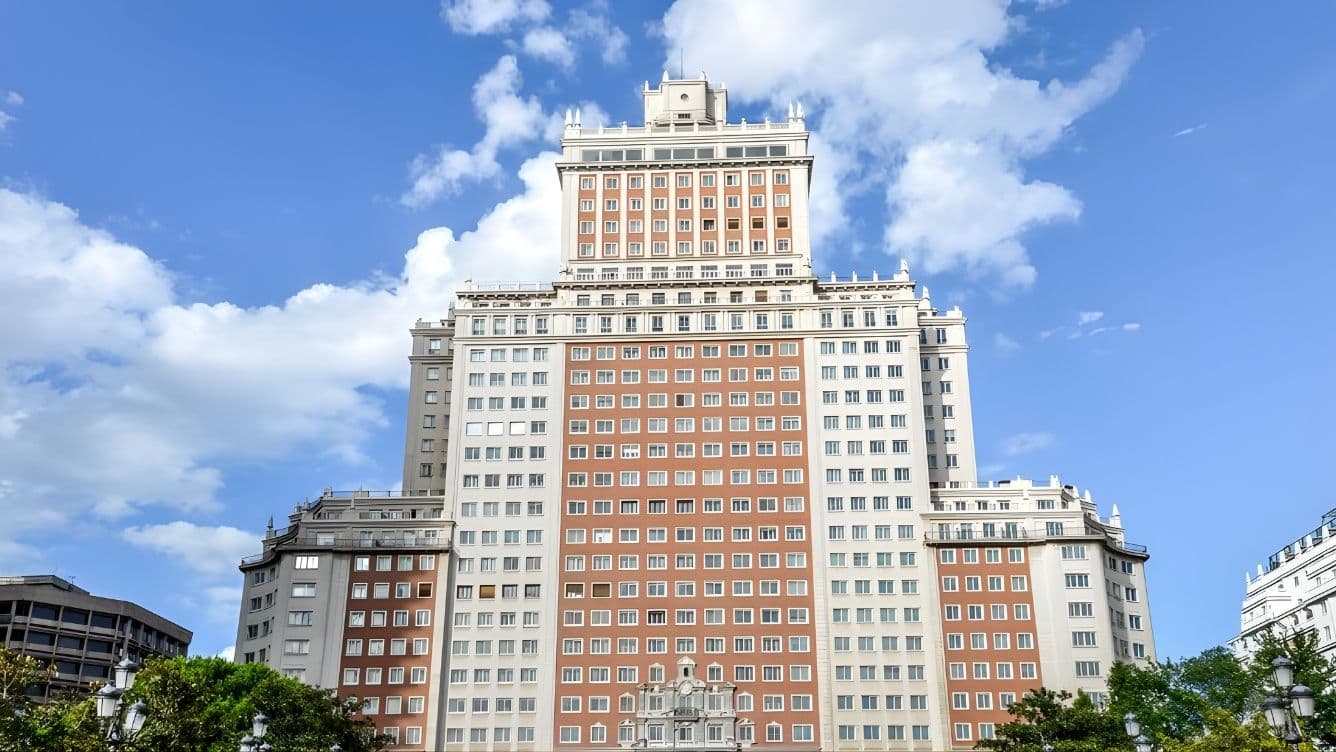 Edifici alt amb múltiples finestres i detalls arquitectònics contra un cel blau amb núvols dispersos. Arbres emmarquen el primer pla.