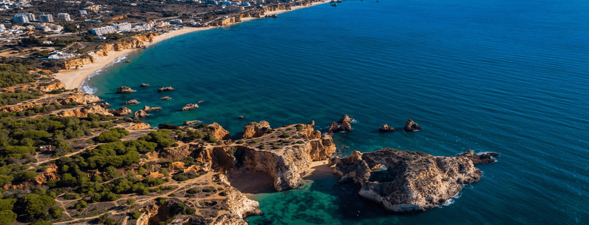 Aerial view of a coastal landscape with rocky cliffs, sandy beaches, turquoise water, and a cityscape in the background under a clear blue sky.