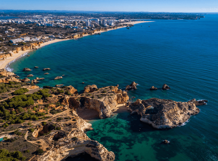 Aerial view of a coastal landscape with rocky cliffs, sandy beaches, turquoise water, and a cityscape in the background under a clear blue sky.