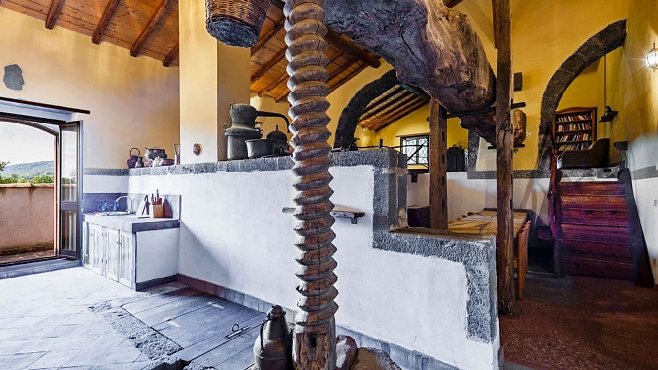 Rustikales Zimmer mit Holzbalken, Steinwänden und einer großen Holzpresse. Die offene Tür gibt den Blick frei auf eine malerische Landschaft. Vintage-Dekor und warme Beleuchtung.