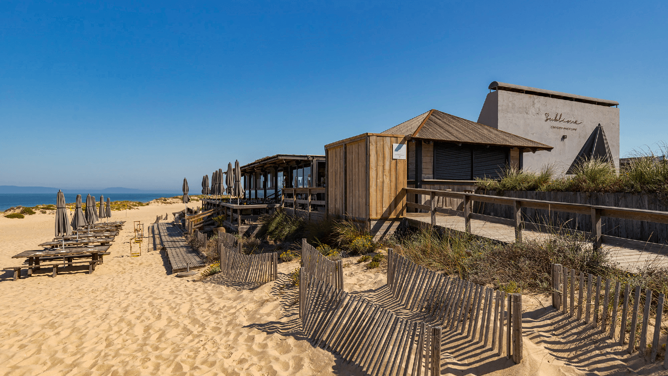 Hölzernes Strandcafé mit geschlossenen Sonnenschirmen, sandigem Pfad und Holzzäunen unter einem klaren blauen Himmel.