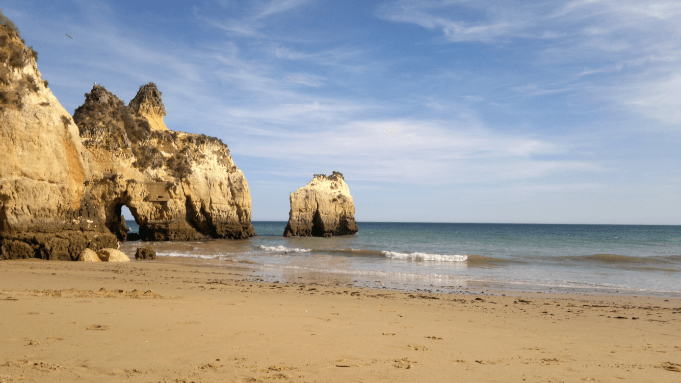 Sandy beach with gentle waves, rocky cliffs, and a clear blue sky in the background.