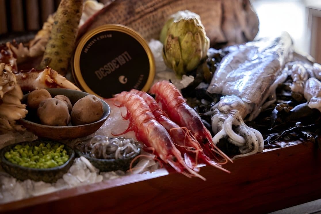 Assorted seafood and vegetables on ice, including red prawns, squid, potatoes, peas, an artichoke, and a small tin can on a wooden tray.