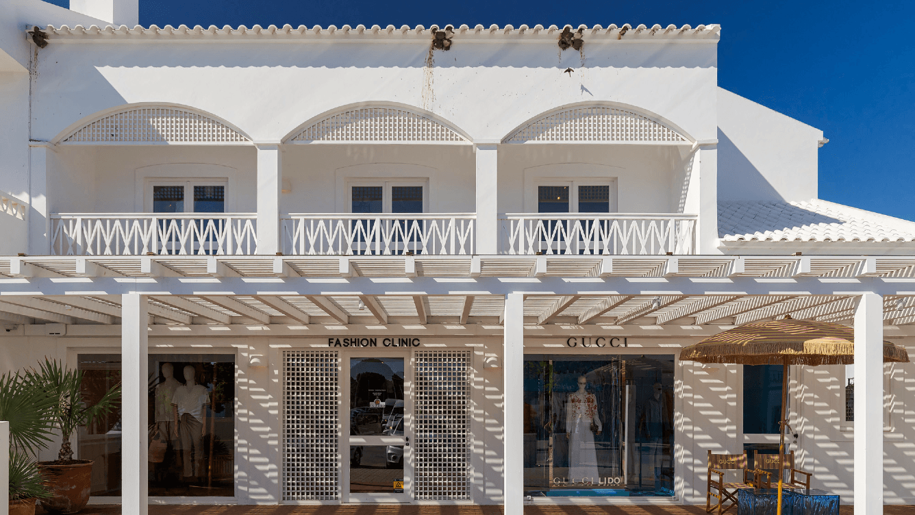 Weißes Gebäude mit Bogenfenstern und zwei Modeboutique-Eingängen darunter, "Fashion Clinic" und "Gucci", unter einer Pergola mit Schatten.