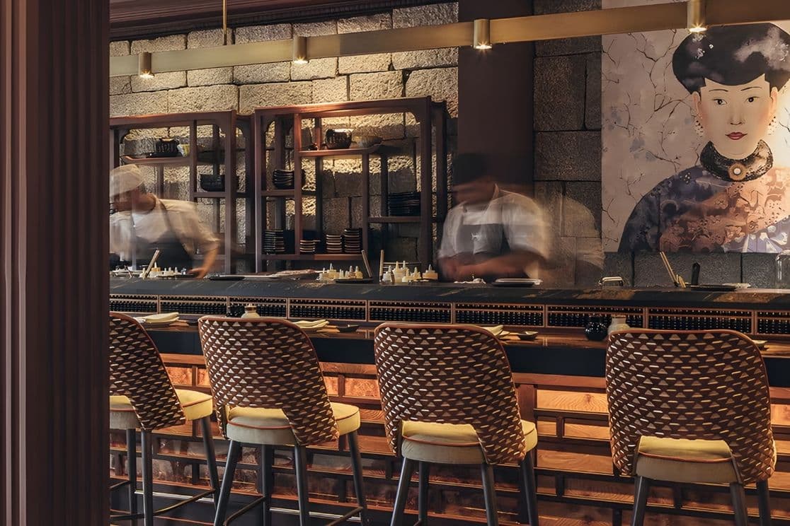 A stylish restaurant interior with chefs preparing food behind a bar, featuring wicker chairs and a large artistic portrait on the wall.