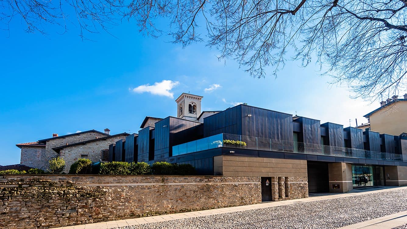 Modernes Gebäude mit dunkler Glasfassade neben historischen Steinstrukturen, unter einem klaren blauen Himmel mit kahlen Ästen darüber.