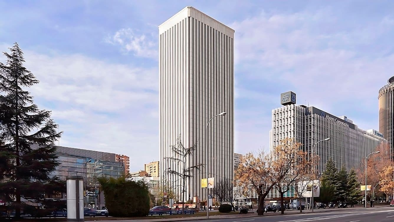 Un gratacel alt i blanc amb línies verticals sota un cel blau clar, envoltat d'altres edificis, arbres i alguns núvols.