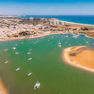 Aerial view of a coastal town with sailboats in a green lagoon, sandy beaches, and a vibrant blue ocean under a clear sky.