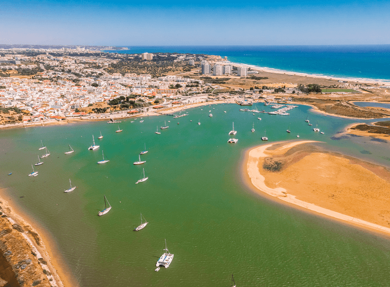 Luftaufnahme einer Küstenstadt mit Segelbooten in einer grünen Lagune, sandigen Stränden und einem lebendigen blauen Ozean unter einem klaren Himmel.