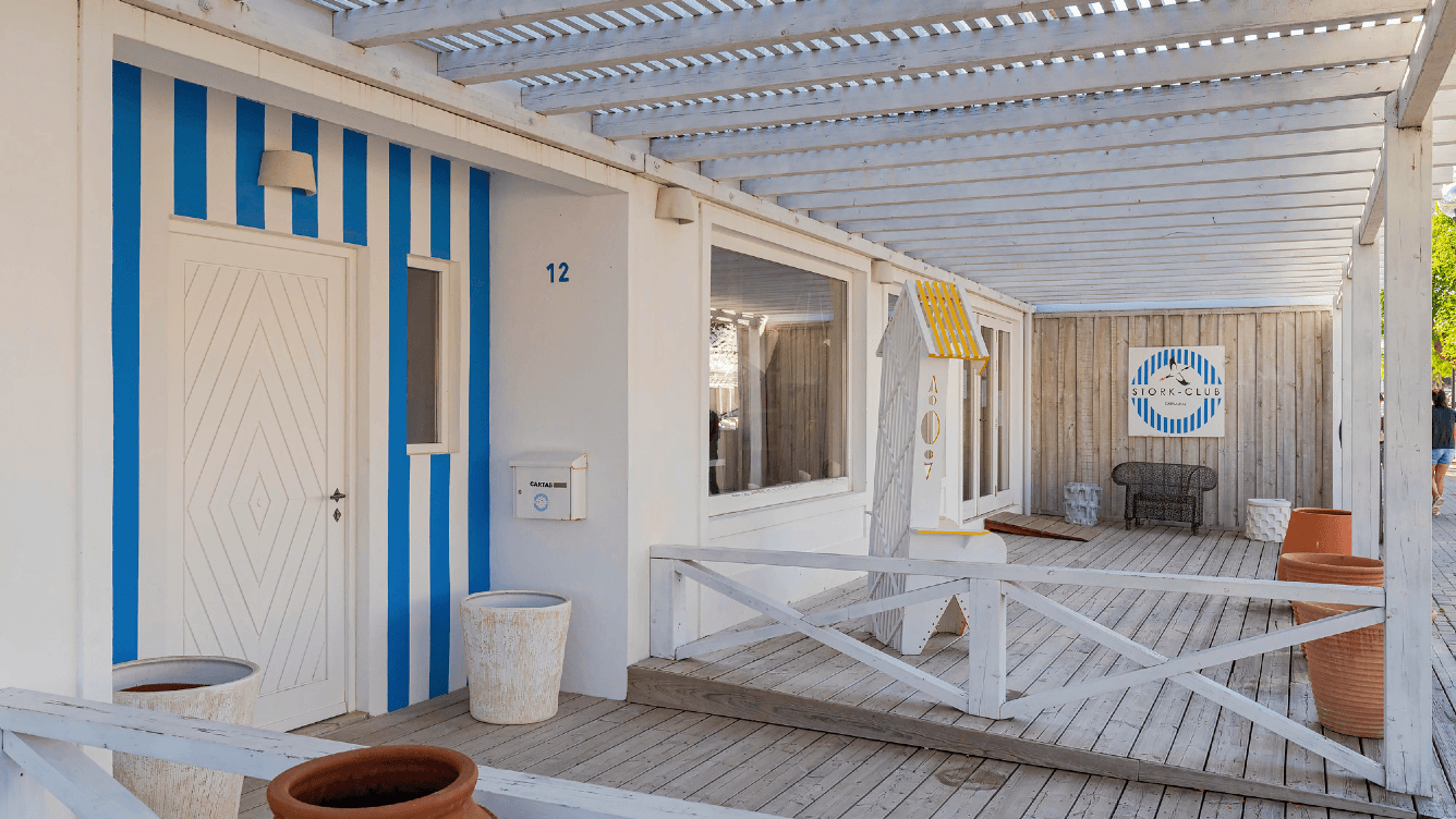 Hölzerner Strandclub-Eingang mit blau-weiß gestreifter Dekoration, weißem Geländer und Topfpflanzen. Ein Stuhl ist in der Ecke sichtbar.