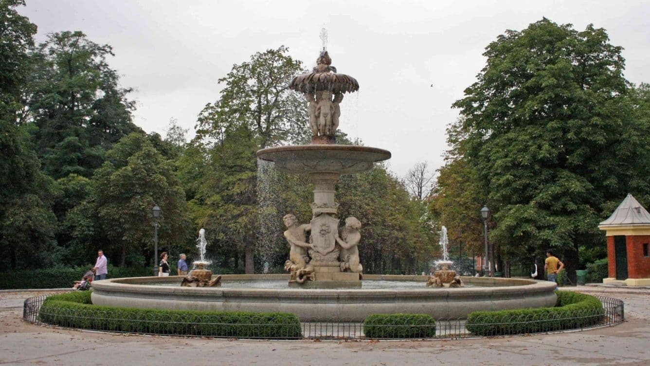 A large ornate fountain with sculptures of cherubs, situated in a park surrounded by lush green trees and a few people walking nearby.