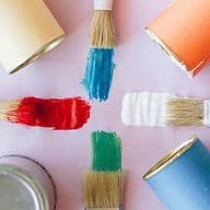 Farbmuster auf einer glatten Oberfläche: Blau, Orange, Gelb und Lila. Es sind vier Pinsel mit roter, blauer, weißer und grüner Farbe sichtbar.