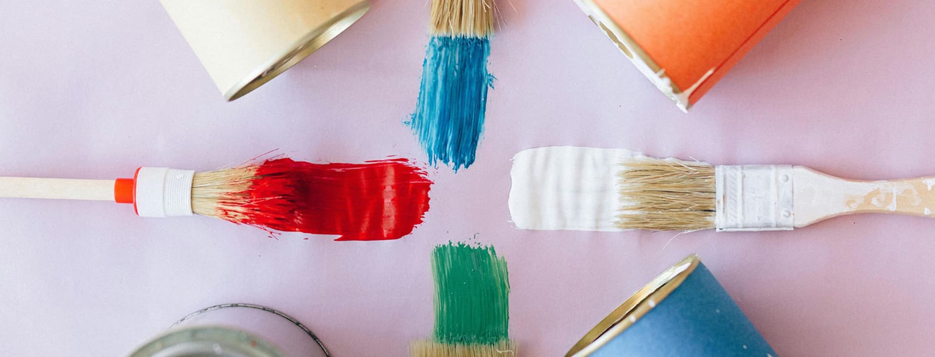 Muestras de pinturas sobre superficie lisa: color azul, naranja, amarillo y lila. Se ven 4 pinceles con color rojo, azul, blanco y verde