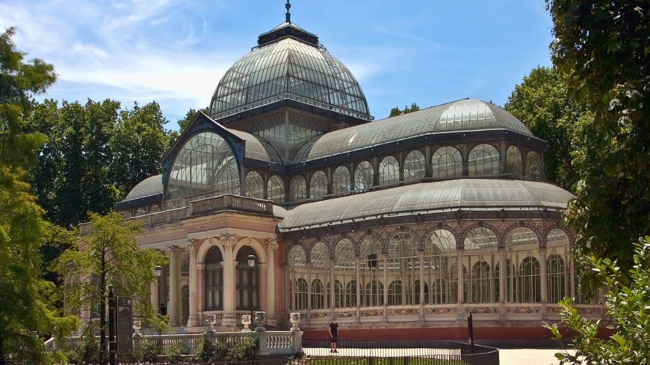 A glass-domed, ornate greenhouse structure surrounded by trees under a bright blue sky.