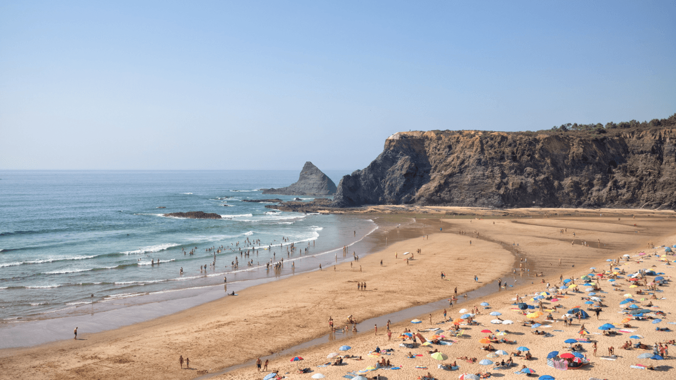 Sunny beach scene with colorful umbrellas, people swimming, and a rocky cliff in the background under a clear blue sky.