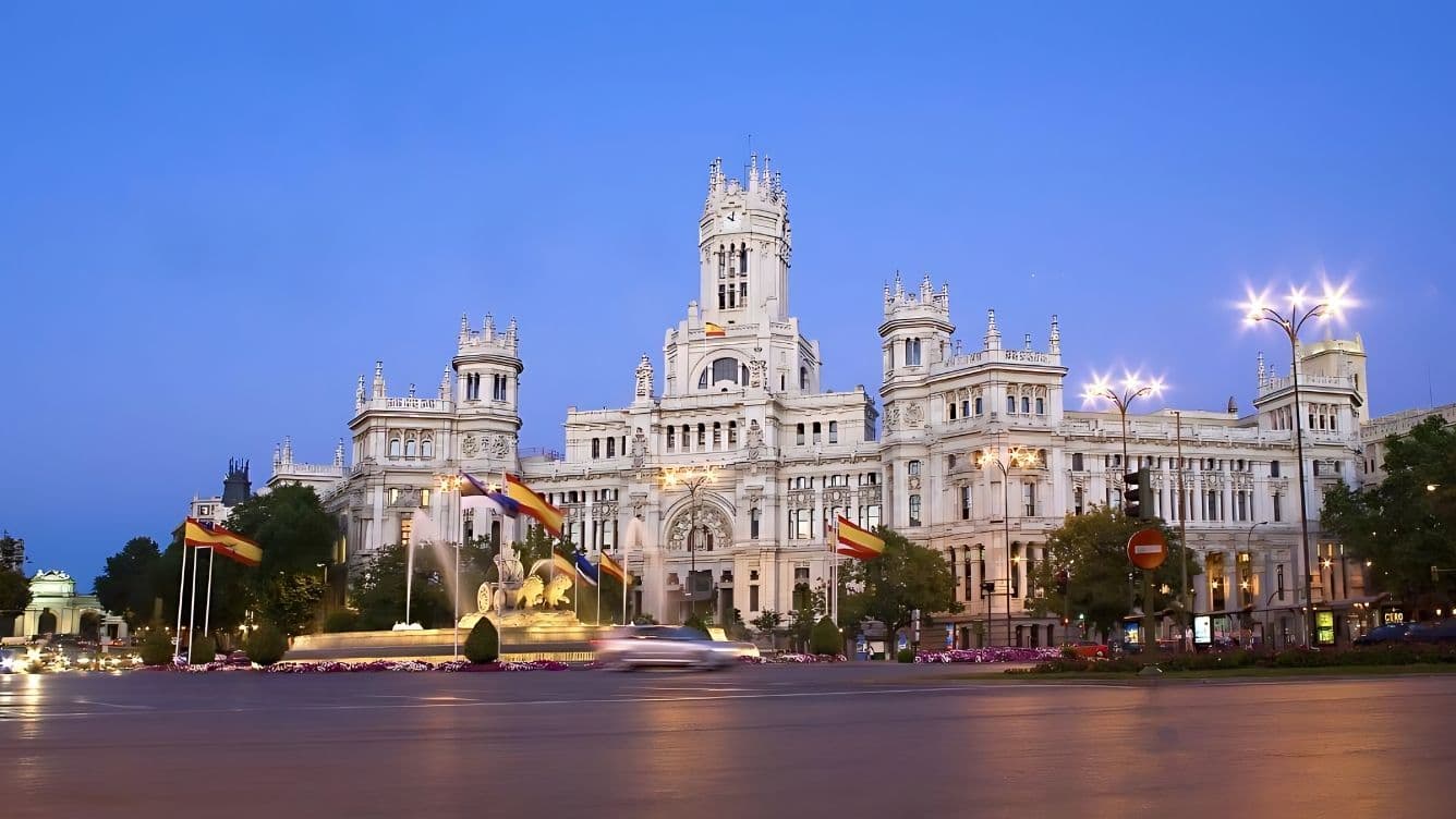 Vista del Palau de Cibeles il·luminat al capvespre a Madrid, amb el trànsit borrós i les banderes espanyoles en primer pla.