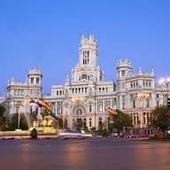 Vista del Palacio de Cibeles iluminado al atardecer en Madrid, con el tráfico borroso y las banderas españolas en primer plano.