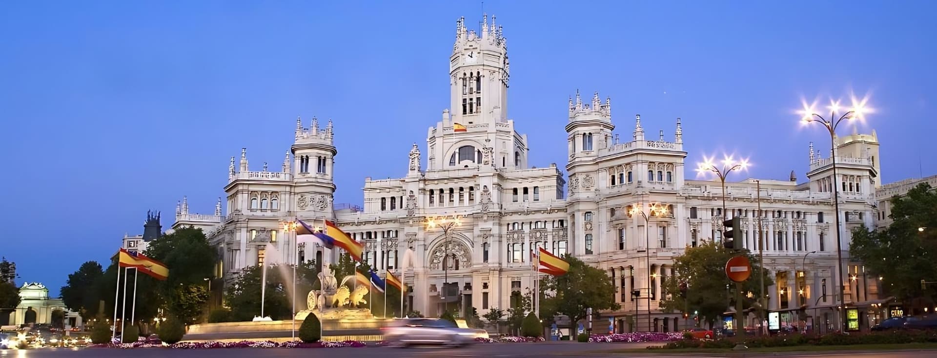 Vista del Palau de Cibeles il·luminat al capvespre a Madrid, amb el trànsit borrós i les banderes espanyoles en primer pla.