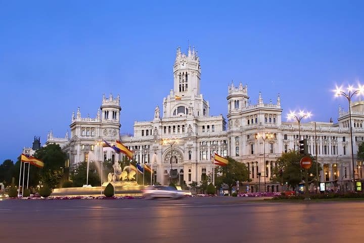 Vista del Palacio de Cibeles iluminado al atardecer en Madrid, con el tráfico borroso y las banderas españolas en primer plano.