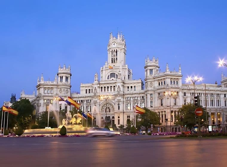 View of the illuminated Palacio de Cibeles at dusk in Madrid, with blurred traffic and Spanish flags in the foreground.
