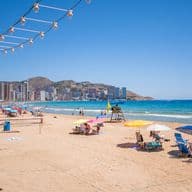 La platja de Llevant de Benidorm, sorra daurada i aigua cristal·lina, gent prenent el sol amb para-sol i gandules.