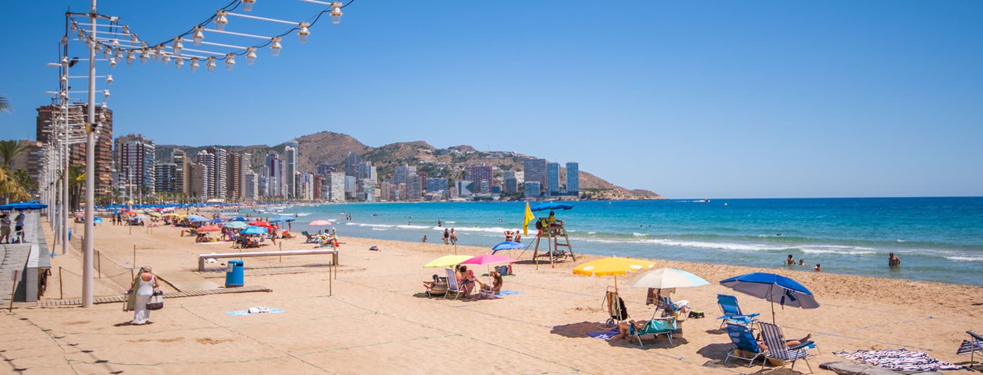 Levante Beach in Benidorm, golden sand and crystal-clear water, people sunbathing with umbrellas and loungers.