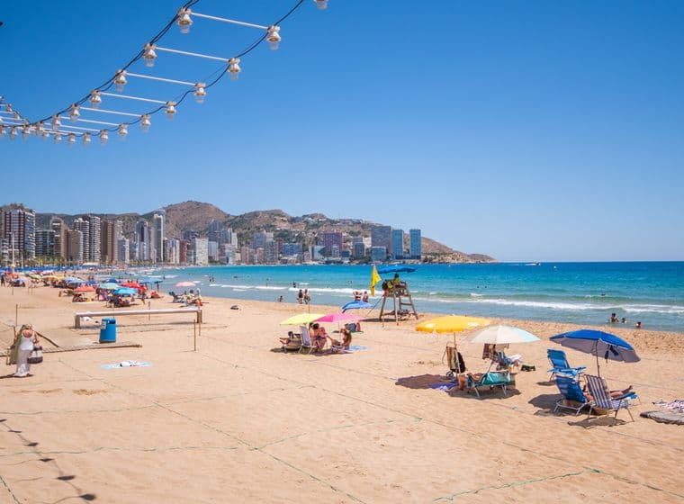 La platja de Llevant de Benidorm, sorra daurada i aigua cristal·lina, gent prenent el sol amb para-sol i gandules.