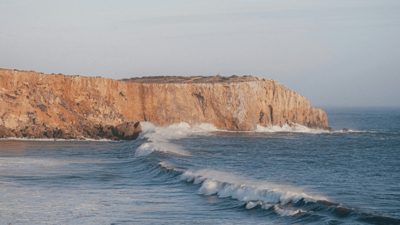 Waves crashing against a rugged cliff under a clear sky, with the ocean extending to the horizon.