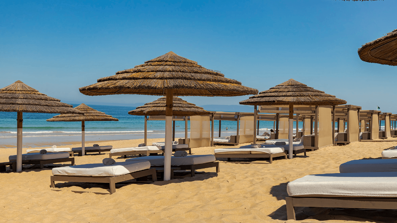 Sandiger Strand mit Strohschirmen und Sonnenliegen unter einem klaren blauen Himmel, mit Blick auf ein ruhiges Meer im Hintergrund.