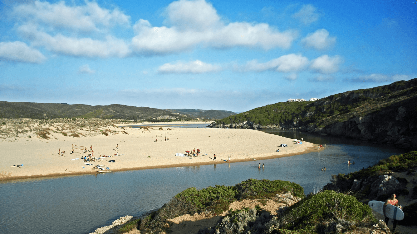 Ein malerischer Strand mit Menschen, die sonnenbaden und in der Nähe eines Flusses schwimmen, umgeben von felsigen Hügeln und Grün unter einem blauen Himmel mit Wolken.