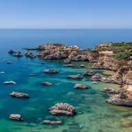 Luftaufnahme einer felsigen Küstenlinie mit türkisfarbenen Gewässern, verstreut liegenden Booten und üppiger Vegetation unter einem klaren blauen Himmel.