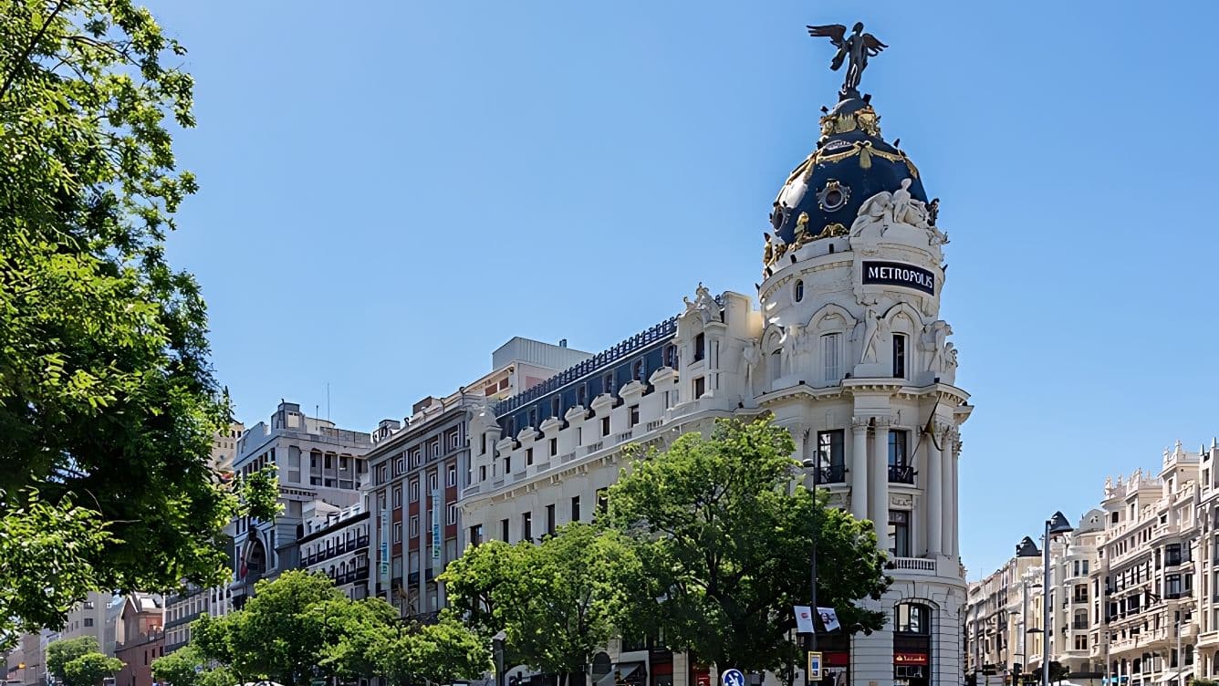 L'edifici Metròpolis de Madrid, amb la seva arquitectura ornamentada, és una cúpula negra coronada per una estàtua alada i envoltat de frondosos arbres.