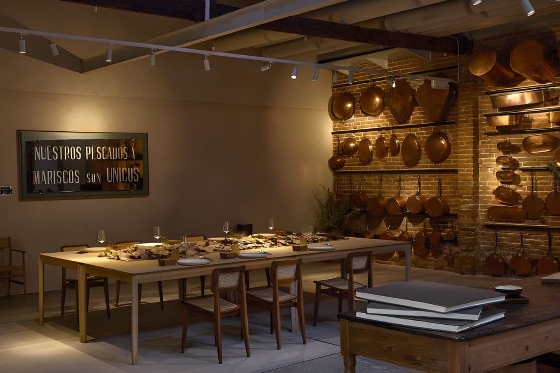 Cozy restaurant with a long wooden table set for dining, warm lighting, and decorative pots on brick walls. Sign reads "Nuestros pescados y mariscos son únicos."