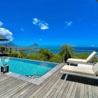 An elegant luxury villa in Mauritius featuring a stunning infinity pool overlooking the turquoise ocean. The wooden deck surrounds the pool with comfortable lounge chairs, while tropical vegetation frames the view of a small island visible in the distance under a bright blue sky with scattered clouds.