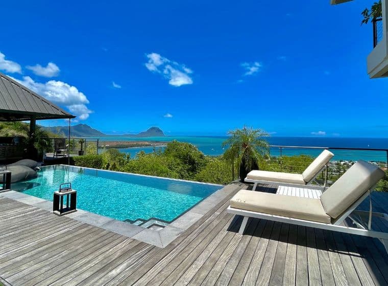 An elegant luxury villa in Mauritius featuring a stunning infinity pool overlooking the turquoise ocean. The wooden deck surrounds the pool with comfortable lounge chairs, while tropical vegetation frames the view of a small island visible in the distance under a bright blue sky with scattered clouds.