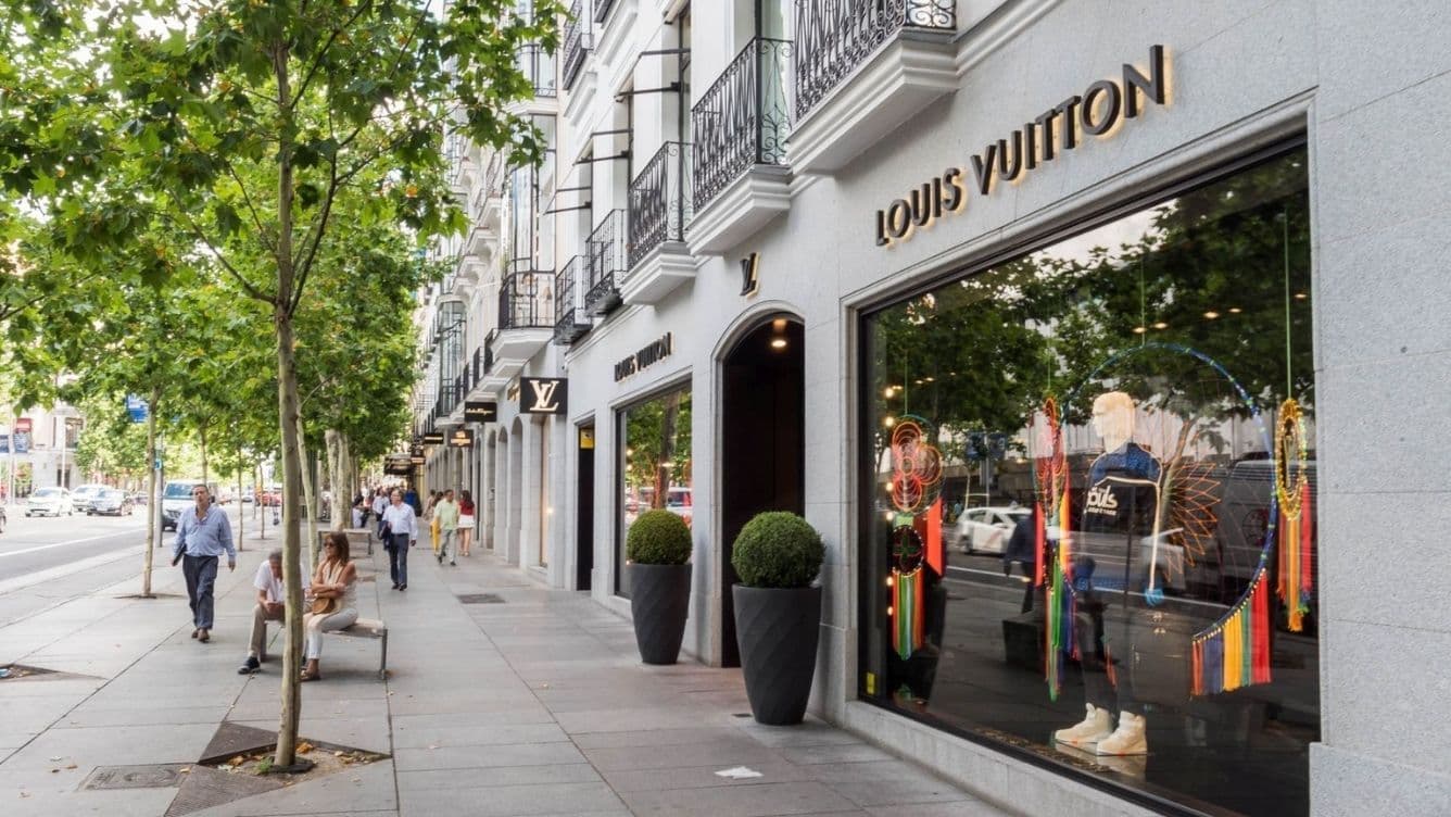 Street view of a luxury storefront with "Louis Vuitton" signage, window displays, and people walking on a tree-lined sidewalk.