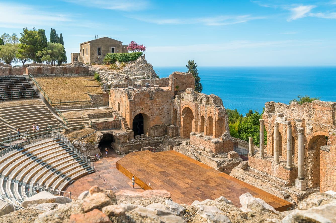 Antikes griechisches Amphitheater mit Meerblick, mit gestuften Sitzplätzen und teilweise zerstörten Bögen, vor einem klaren blauen Himmel.