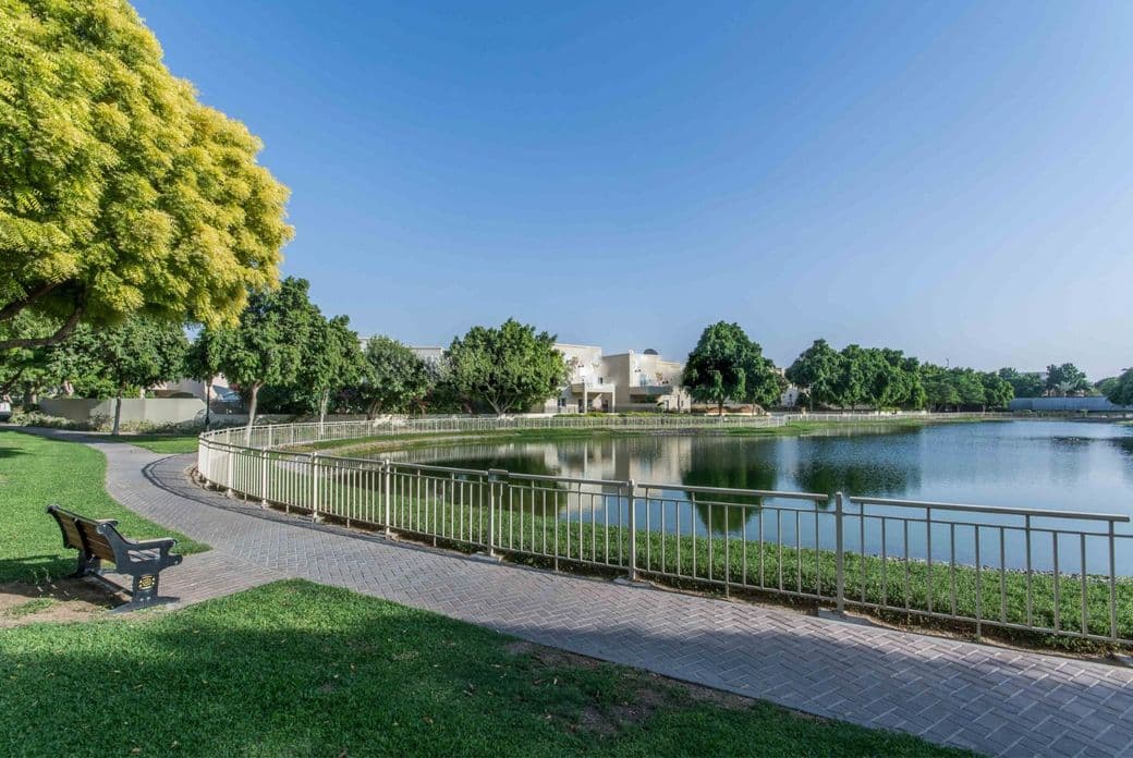 A serene park scene in The Lakes community with a curved path, a bench, a pond, and lush green trees under a clear blue sky.