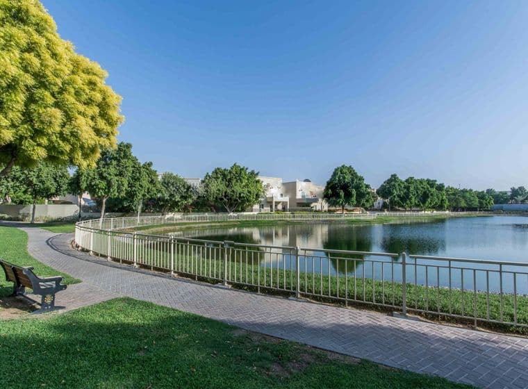 A serene park scene in The Lakes community with a curved path, a bench, a pond, and lush green trees under a clear blue sky.