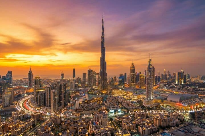 Aerial view of Downtown Dubai at sunset, featuring Burj Khalifa surrounded by illuminated buildings and a winding highway.