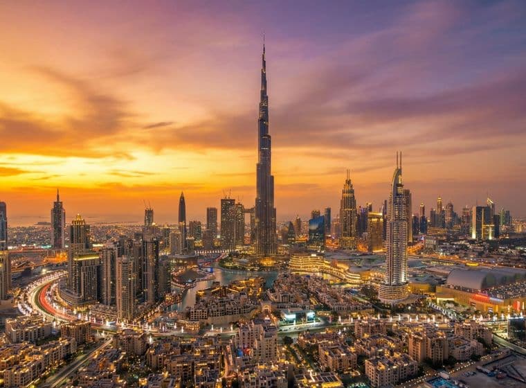 Aerial view of Downtown Dubai at sunset, featuring Burj Khalifa surrounded by illuminated buildings and a winding highway.
